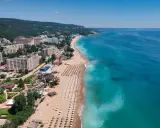 Słoneczny dzień nad morzem, gdzie złote piaski plaży wylegują się w słońcu, a turkusowa woda zaprasza do kąpieli. W tle hotelowe budynki i zielone wzgórza.