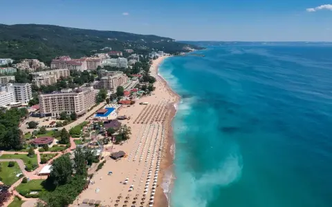Słoneczny dzień nad morzem, gdzie złote piaski plaży wylegują się w słońcu, a turkusowa woda zaprasza do kąpieli. W tle hotelowe budynki i zielone wzgórza.