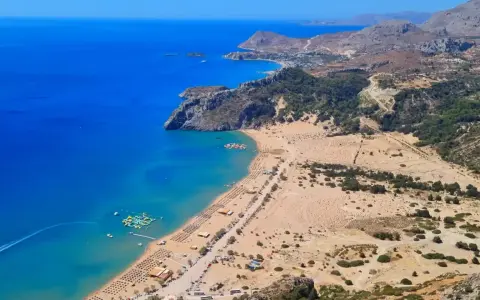Rodos zachwyca turkusowym morzem i piaszczystą plażą. W oddali widać góry i zabudowania. Idealne miejsce na wakacje.