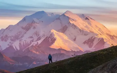 Widok na najwyższy szczyt Ameryki Północnej, Denali, oświetlony zachodzącym słońcem. Samotny turysta stoi na zboczu, podziwiając majestat góry.