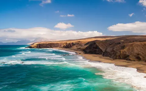 Fuerteventura w lutym: turkusowe fale rozbijają się o piaszczystą plażę, a za nią wznoszą się surowe, brązowe klify.