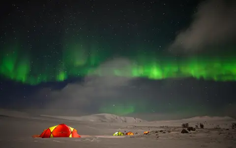 Nocne niebo rozświetla zorza polarna, a pod nią rozbijają się namioty. Idealny moment na obserwację tego zjawiska.