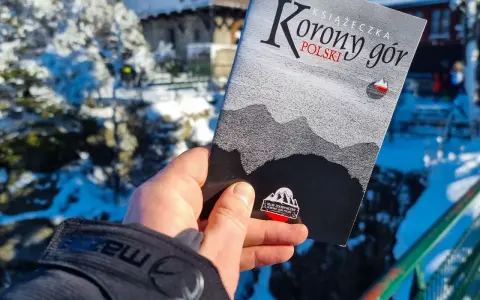 Dłoń trzyma książeczkę "Korona Gór Polski" na tle zimowego krajobrazu.