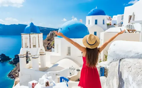 Santorini, Grecja, gdzie warto pojechać. Kobieta w czerwonej sukience i kapeluszu cieszy się widokiem białych domów i niebieskich kopuł nad morzem.